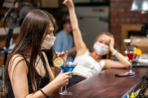 girls in medical masks chatting in a pub. girls in medical masks are talking at the bar. girls in medical masks drink cocktails