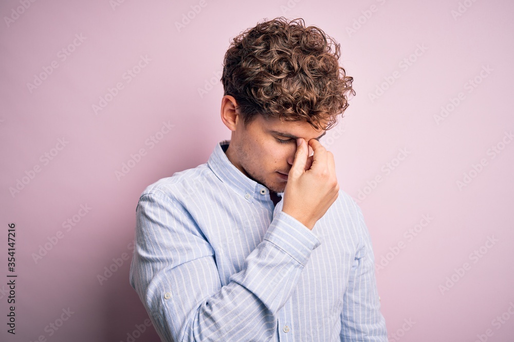 Young blond handsome man with curly hair wearing striped shirt over white background tired rubbing nose and eyes feeling fatigue and headache. Stress and frustration concept. Young blond handsome man with curly hair wearing striped shirt over white background tired rubbing nose and eyes feeling fatigue and headache. Stress and frustration concept.