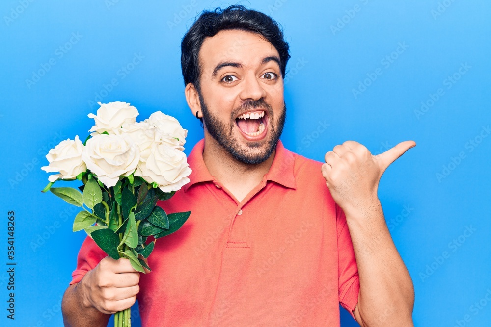 Young hispanic man holding flowers pointing thumb up to the side smiling happy with open mouth Young hispanic man holding flowers pointing thumb up to the side smiling happy with open mouth