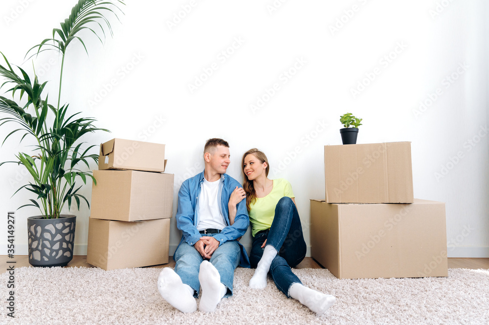 Relocation into new home. Happy guy and girl loving each other moving in a new home, they are sitting on the floor in casual wear and hugging among cardboard boxes Relocation into new home. Happy guy and girl loving each other moving in a new home, they are sitting on the floor in casual wear and hugging among cardboard boxes