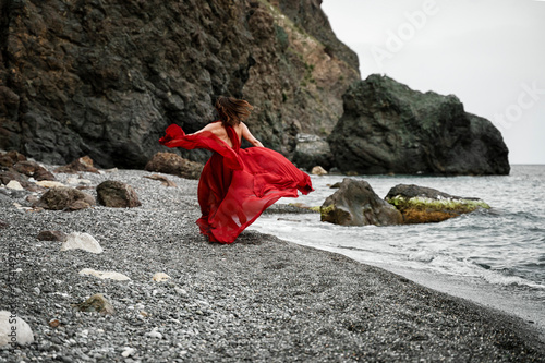 A beautiful girl in a long red dress that flutters in the wind, runs along the beach with the concept of femininity, harmony. Black rocks and rocks on the shore.