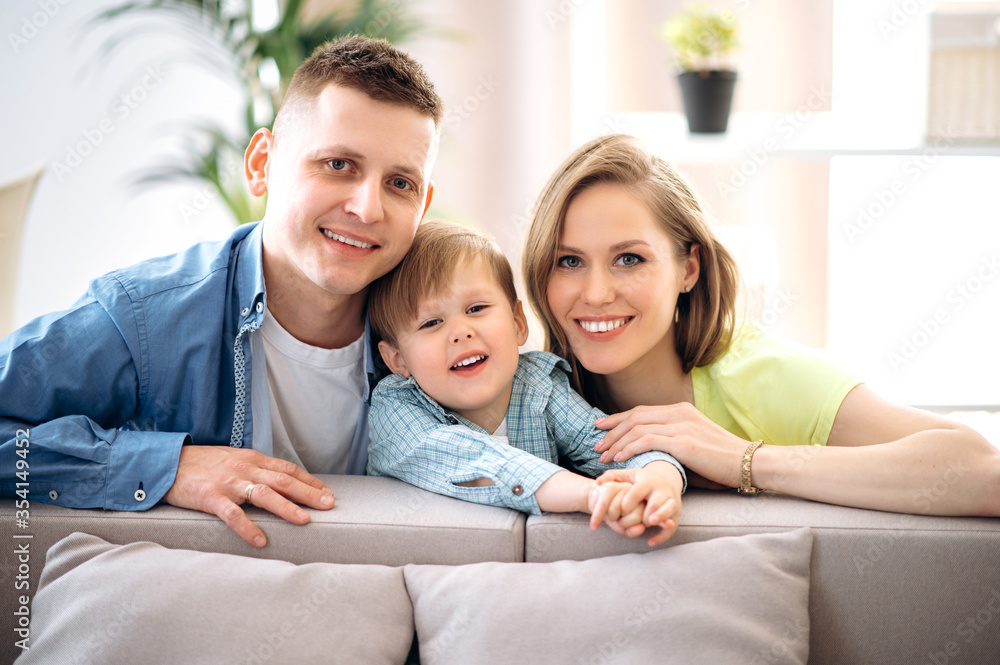 Fototapeta premium Family portrait. A happy family. Handsome dad, attractive mom and their little adorable smiling son are sitting on the sofa in their cozy apartment