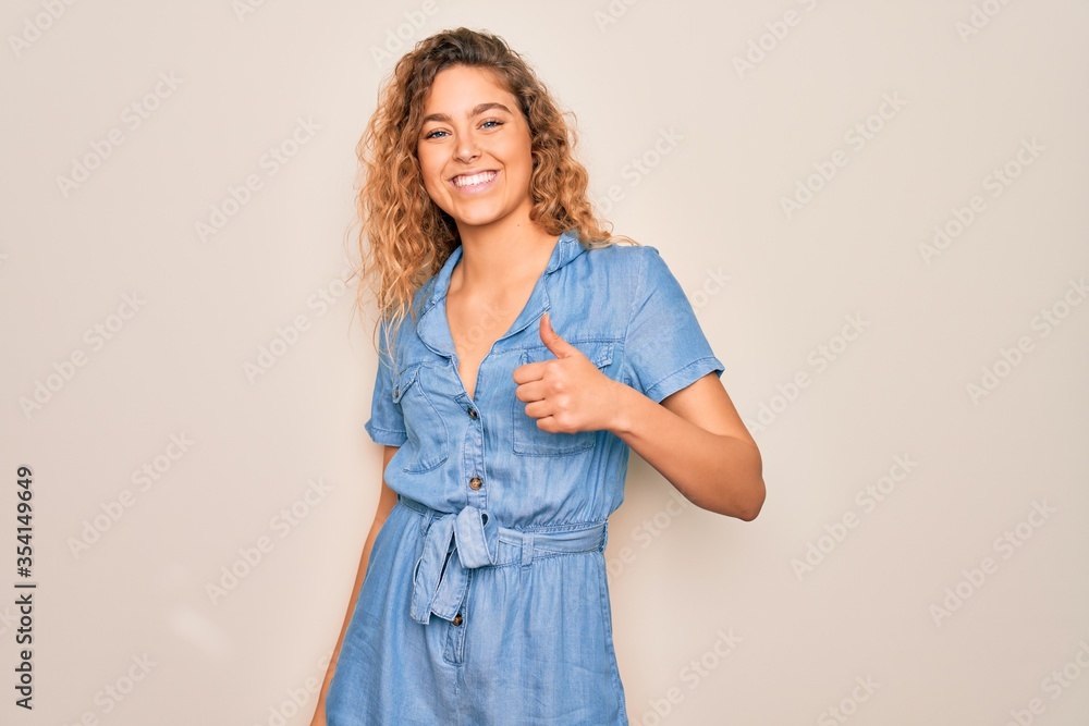 Young beautiful woman with blue eyes wearing casual denim dress over white background doing happy thumbs up gesture with hand. Approving expression looking at the camera showing success. Young beautiful woman with blue eyes wearing casual denim dress over white background doing happy thumbs up gesture with hand. Approving expression looking at the camera showing success.