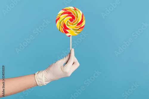 Profile side view closeup of human hand in white surgical gloves holding and showing rainbow colorful candy in hand. indoor, studio shot, isolated on blue background.