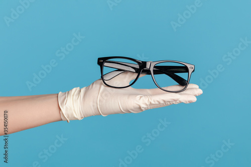 Profile side view closeup of human hand in white surgical gloves holding and giving black eyeglasses frame. indoor, studio shot, isolated on blue background.