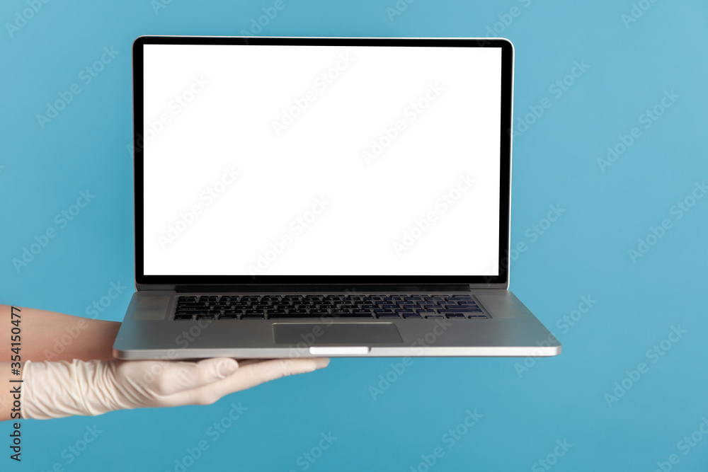 Profile side view closeup of human hand in white surgical gloves holding computer laptop and showing empty copy space on monitor screen. indoor, studio shot, isolated on blue background. Profile side view closeup of human hand in white surgical gloves holding computer laptop and showing empty copy space on monitor screen. indoor, studio shot, isolated on blue background.