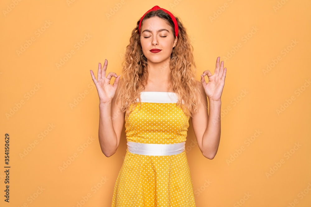 Beautiful blonde pin-up woman with blue eyes wearing diadem standing over yellow background relaxed and smiling with eyes closed doing meditation gesture with fingers. Yoga concept. Beautiful blonde pin-up woman with blue eyes wearing diadem standing over yellow background relaxed and smiling with eyes closed doing meditation gesture with fingers. Yoga concept.