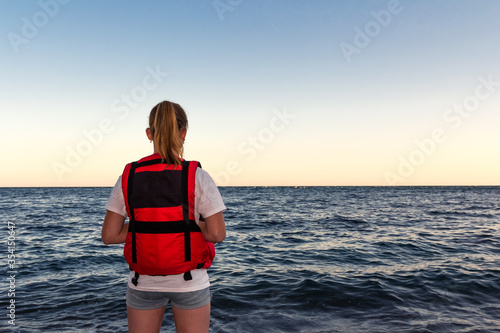 Photography Girl in bright red life jacket stands on seashore with waves, rear view