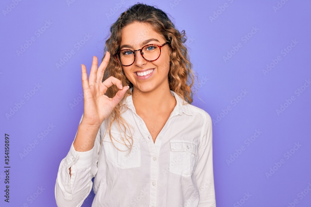 Young beautiful woman with blue eyes wearing casual shirt and glasses over purple background smiling positive doing ok sign with hand and fingers. Successful expression. Young beautiful woman with blue eyes wearing casual shirt and glasses over purple background smiling positive doing ok sign with hand and fingers. Successful expression.