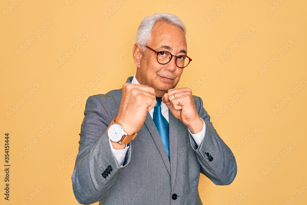 Middle age senior grey-haired handsome business man wearing glasses over yellow background Ready to fight with fist defense gesture, angry and upset face, afraid of problem Middle age senior grey-haired handsome business man wearing glasses over yellow background Ready to fight with fist defense gesture, angry and upset face, afraid of problem