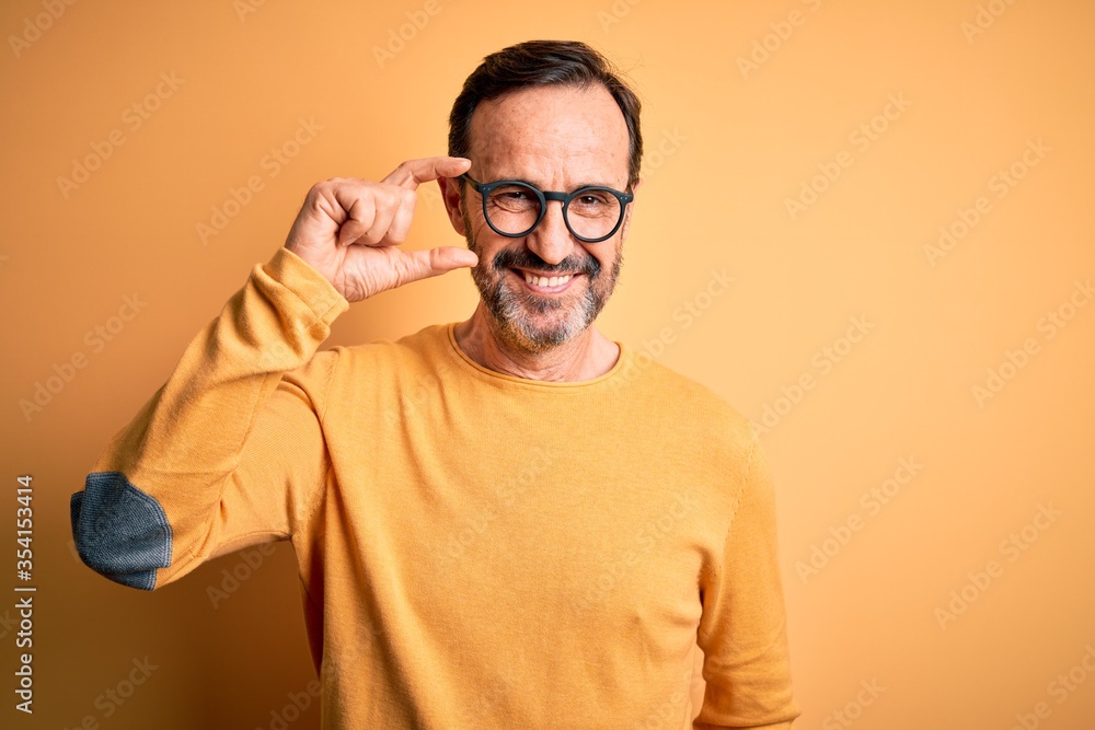 Middle age hoary man wearing casual sweater and glasses over isolated yellow background smiling and confident gesturing with hand doing small size sign with fingers looking and the camera. Measure Middle age hoary man wearing casual sweater and glasses over isolated yellow background smiling and confident gesturing with hand doing small size sign with fingers looking and the camera. Measure
