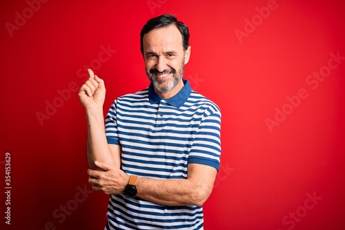 Middle age hoary man wearing casual striped polo standing over isolated red background with a big smile on face, pointing with hand and finger to the side looking at the camera.