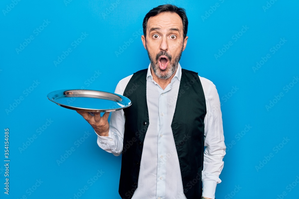 Middle age handsome waitress man holding tray standing over isolated blue background scared and amazed with open mouth for surprise, disbelief face Middle age handsome waitress man holding tray standing over isolated blue background scared and amazed with open mouth for surprise, disbelief face