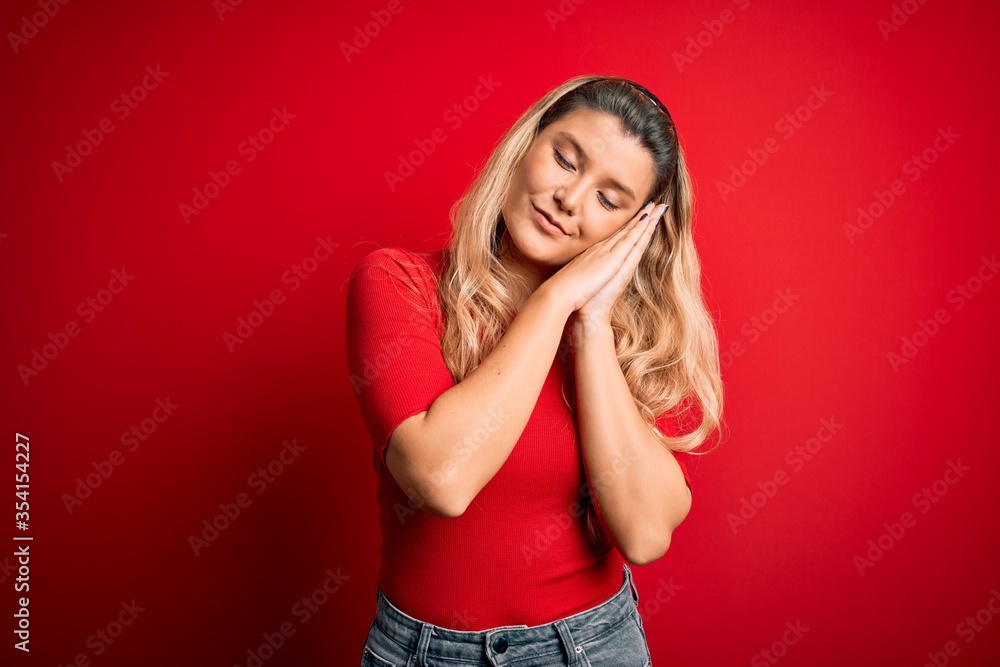 Young beautiful blonde woman wearing casual t-shirt standing over isolated red background sleeping tired dreaming and posing with hands together while smiling with closed eyes. Young beautiful blonde woman wearing casual t-shirt standing over isolated red background sleeping tired dreaming and posing with hands together while smiling with closed eyes.