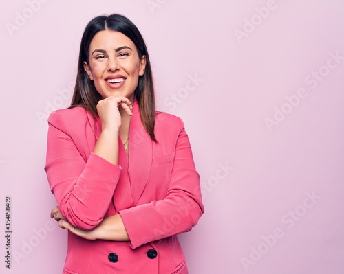 Young beautiful business woman wearing elegant jacket standing over pink background smiling looking confident at the camera with crossed arms and hand on chin. Thinking positive.