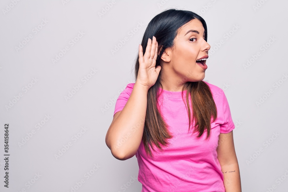 Young beautiful brunette woman wearing casual pink t-shirt standing over white background smiling with hand over ear listening an hearing to rumor or gossip. Deafness concept. Young beautiful brunette woman wearing casual pink t-shirt standing over white background smiling with hand over ear listening an hearing to rumor or gossip. Deafness concept.