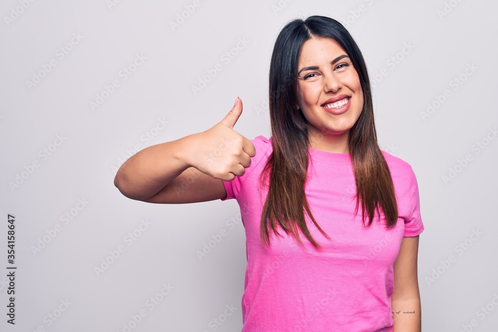 Young beautiful brunette woman wearing casual pink t-shirt standing over white background doing happy thumbs up gesture with hand. Approving expression looking at the camera showing success. Young beautiful brunette woman wearing casual pink t-shirt standing over white background doing happy thumbs up gesture with hand. Approving expression looking at the camera showing success.