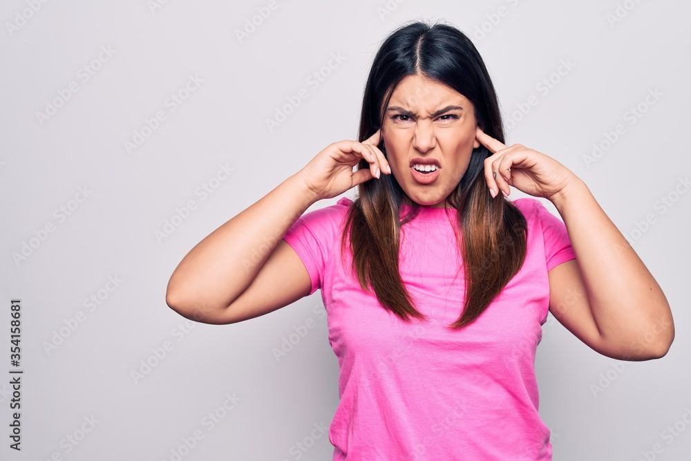 Young beautiful brunette woman wearing casual pink t-shirt standing over white background covering ears with fingers with annoyed expression for the noise of loud music. Deaf concept. Young beautiful brunette woman wearing casual pink t-shirt standing over white background covering ears with fingers with annoyed expression for the noise of loud music. Deaf concept.