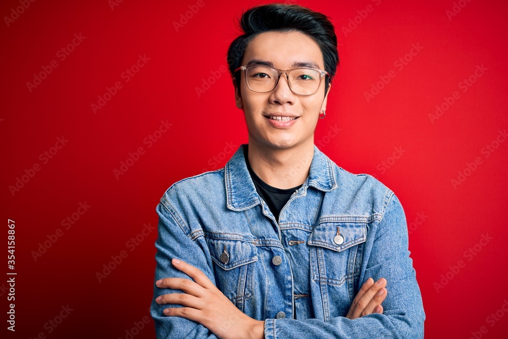 Young handsome chinese man wearing denim jacket and glasses over red background happy face smiling with crossed arms looking at the camera. Positive person. Young handsome chinese man wearing denim jacket and glasses over red background happy face smiling with crossed arms looking at the camera. Positive person.