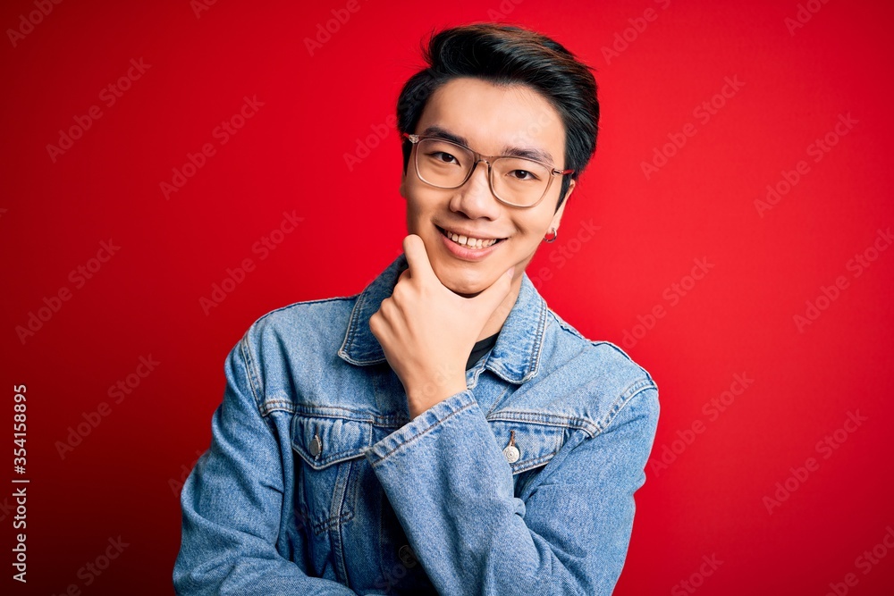 Obraz premium Young handsome chinese man wearing denim jacket and glasses over red background looking confident at the camera smiling with crossed arms and hand raised on chin. Thinking positive.
