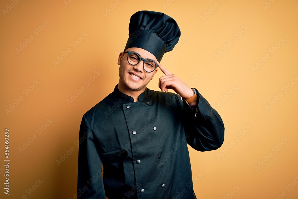 Young brazilian chef man wearing cooker uniform and hat over isolated yellow background Smiling pointing to head with one finger, great idea or thought, good memory Young brazilian chef man wearing cooker uniform and hat over isolated yellow background Smiling pointing to head with one finger, great idea or thought, good memory