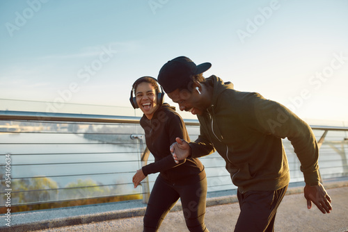 Enjoying run. Side view of happy young African couple, man and woman in headphones laughing while running on the bridge in the early morning