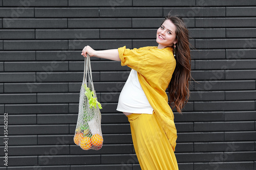 Pregnant woman in yellow clothes with an eco bag with fruits and vegetables on a black wall background. Plastic free concept photo