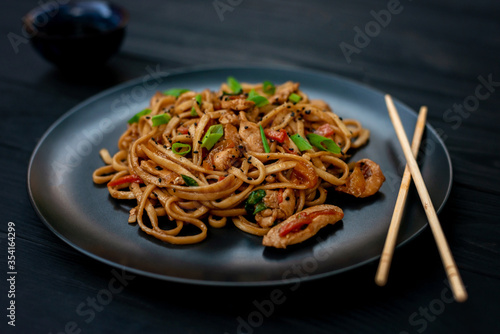 Asian udon noodles with chicken vegetables and teriyaki sauce with chopsticks on a black wooden background. Chinese and Japanese cuisine.
