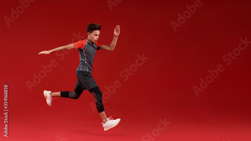 Just run. A teenage boy is engaged in sport, he is looking down while jumping. Isolated on red background. Fitness, training, active lifestyle concept. Horizontal shot