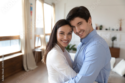 Portrait of happy young family renters stand in living room hug and embrace excited to move together, smiling caucasian couple cuddle show love and affection, enjoy tender romantic weekend at home