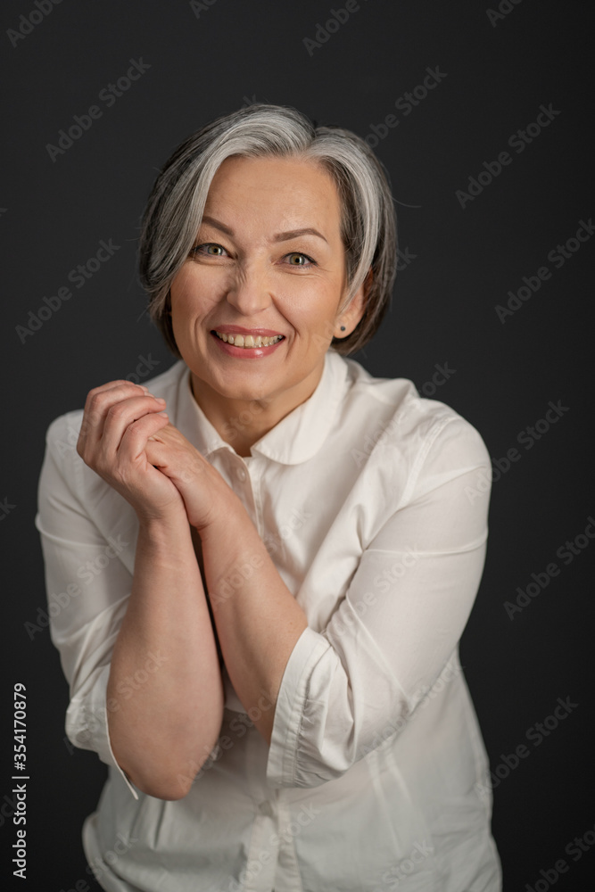 Joyful Caucasian woman smiles broadly while looking at camera and clasped her hands. Enthusiastic gray-haired mature woman in a white shirt posing in studio. Joyful Caucasian woman smiles broadly while looking at camera and clasped her hands. Enthusiastic gray-haired mature woman in a white shirt posing in studio.