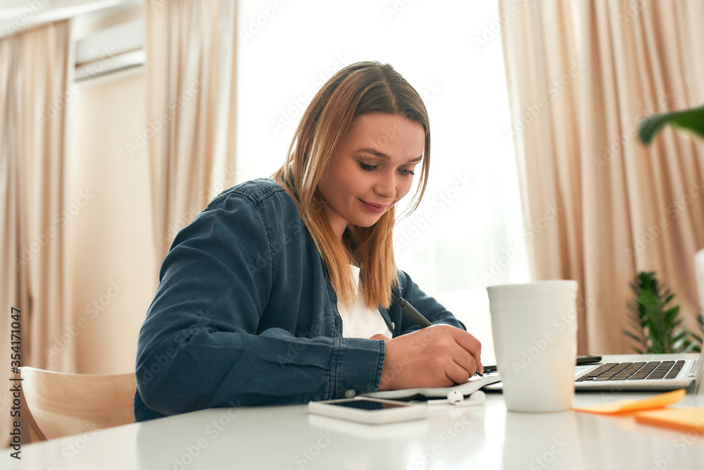 Planning project. Young beautiful female freelancer in casual clothes sitting at her workplace at home and making some notes. Woman working from home Planning project. Young beautiful female freelancer in casual clothes sitting at her workplace at home and making some notes. Woman working from home