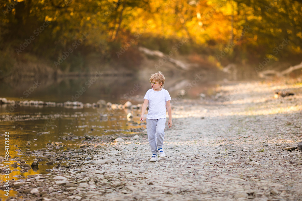 Little blonde boy in green glasses playing on the riverside. Autumn in the yellow forest Little blonde boy in green glasses playing on the riverside. Autumn in the yellow forest