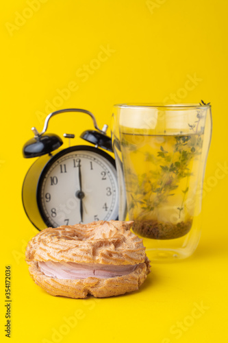 Minimalist stylish food photoshoot in a studio: choux ring with custard cream, herbal tea and black analog clock on yellow background