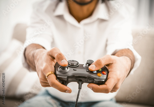 Fair-skinned guy shakes gamepad buttons. Young man in white shirt using wired gamepad while playing computer game. Playing guy concept. Close up shot. Toned image.