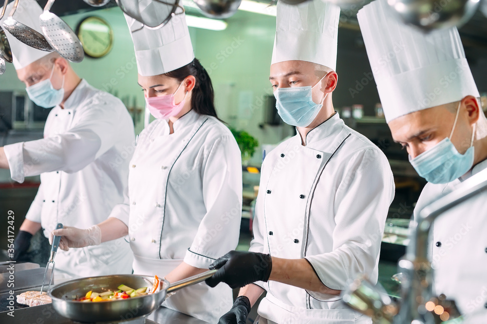 Chefs in protective masks and gloves prepare food in the kitchen of a restaurant or hotel. Chefs in protective masks and gloves prepare food in the kitchen of a restaurant or hotel.