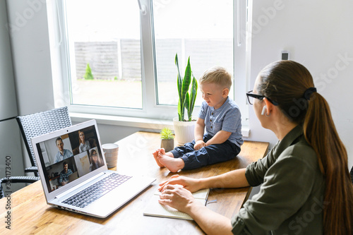 Stay at home. Mother working from home, she talks online with a coworkers via video call on laptop, a cute kid near