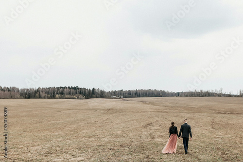 Fototapeta Naklejka Na Ścianę i Meble -  a young couple holding hands goes into the distance along a yellow autumn field towards the forest