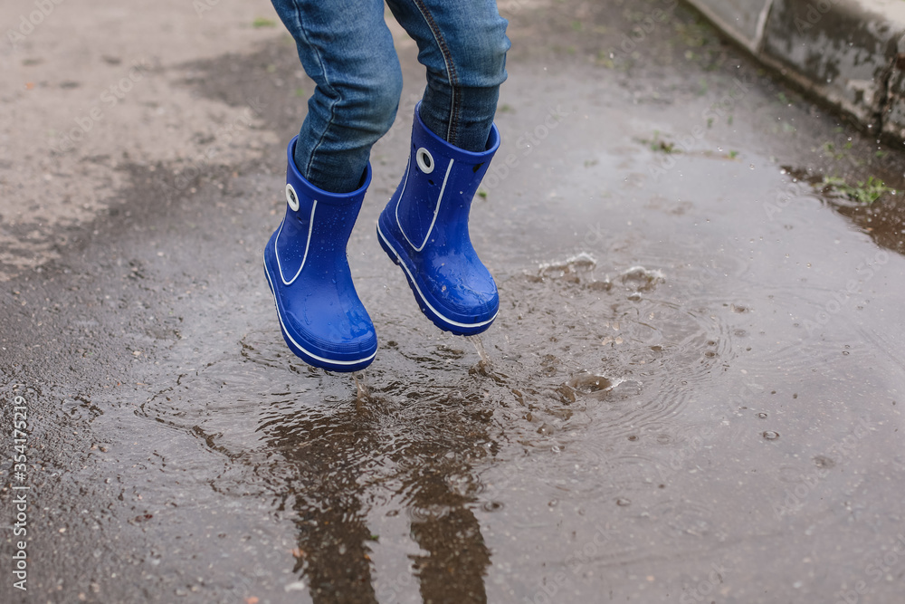 A little boy in blue rubber boots jumps into a puddle. Сlose-up of feet.