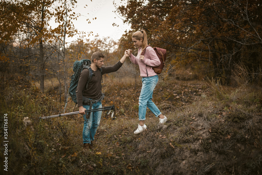 Fototapeta premium Hiking in nature. Young Man and Woman backpackers descend from the slope holding hands and holding hiking poles in their hands. Tourists with backpacks walk through the autumn forest. Support concept.