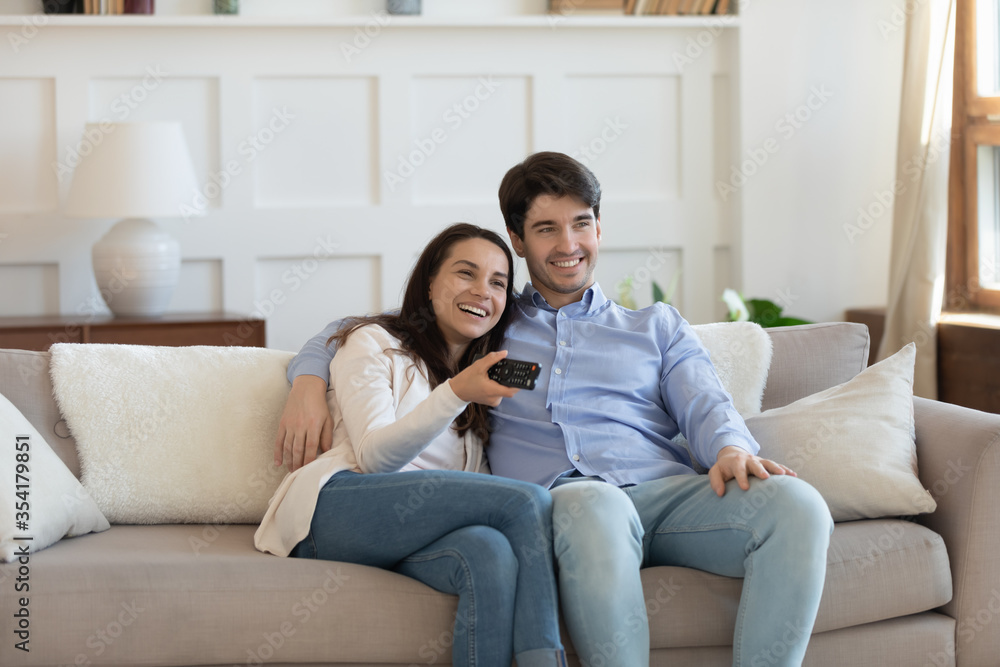 Overjoyed young caucasian couple sit relax on comfortable sofa in living room watching TV together, happy millennial man and woman rest on couch at home enjoy television program on leisure weekend Overjoyed young caucasian couple sit relax on comfortable sofa in living room watching TV together, happy millennial man and woman rest on couch at home enjoy television program on leisure weekend