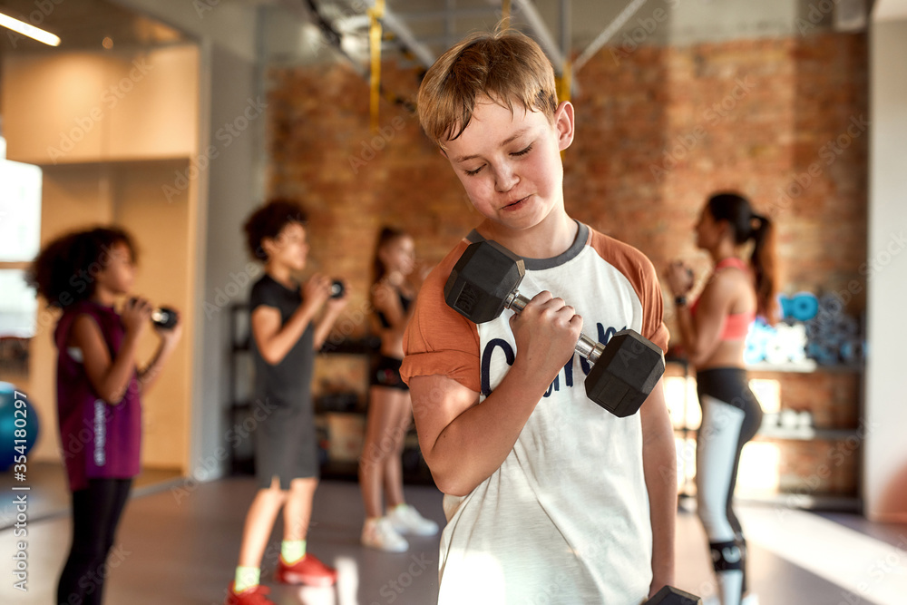 Caucasian boy smiling while exercising using dumbbell in gym together with female trainer and other kids. Sport, healthy lifestyle, physical education concept Caucasian boy smiling while exercising using dumbbell in gym together with female trainer and other kids. Sport, healthy lifestyle, physical education concept