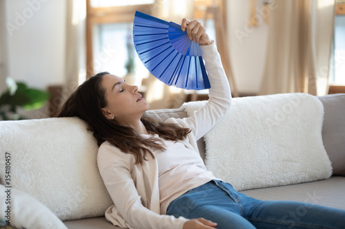 Exhausted young caucasian woman sit relax on couch in living room wave with hand fan suffer from hot weather, overheated female use waver feel unwell unhealthy lack air condition system indoors