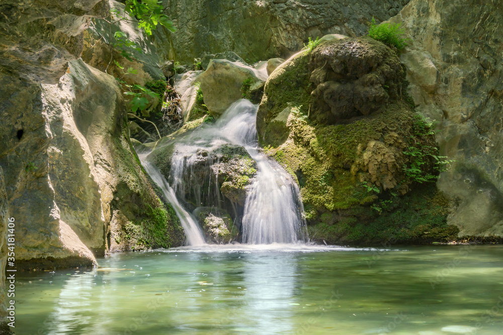Fototapeta premium Beautiful cascade waterfall between rocks covered with moss and plants, small lake at the bottom. Long exposure shot, motion photo.