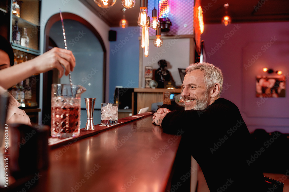 Having a drink. Cheerful handsome middle-aged man sitting at bar counter while bartender making a cocktail for him