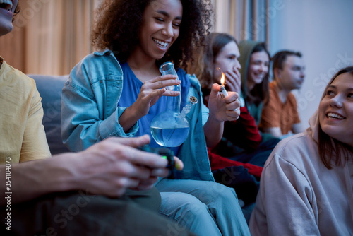 Friends smoking weed at home. Young afro american girl lighting marijuana in the glass bong, relaxing with friends on the sofa at home. Young people playing video games