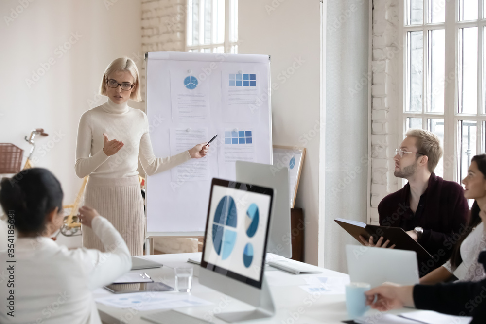 Young serious blonde female trainer coach mentor giving education lecture to motivated diverse employees in modern office. Millennial speaker in eyeglasses explaining marketing strategy to colleagues. Young serious blonde female trainer coach mentor giving education lecture to motivated diverse employees in modern office. Millennial speaker in eyeglasses explaining marketing strategy to colleagues.