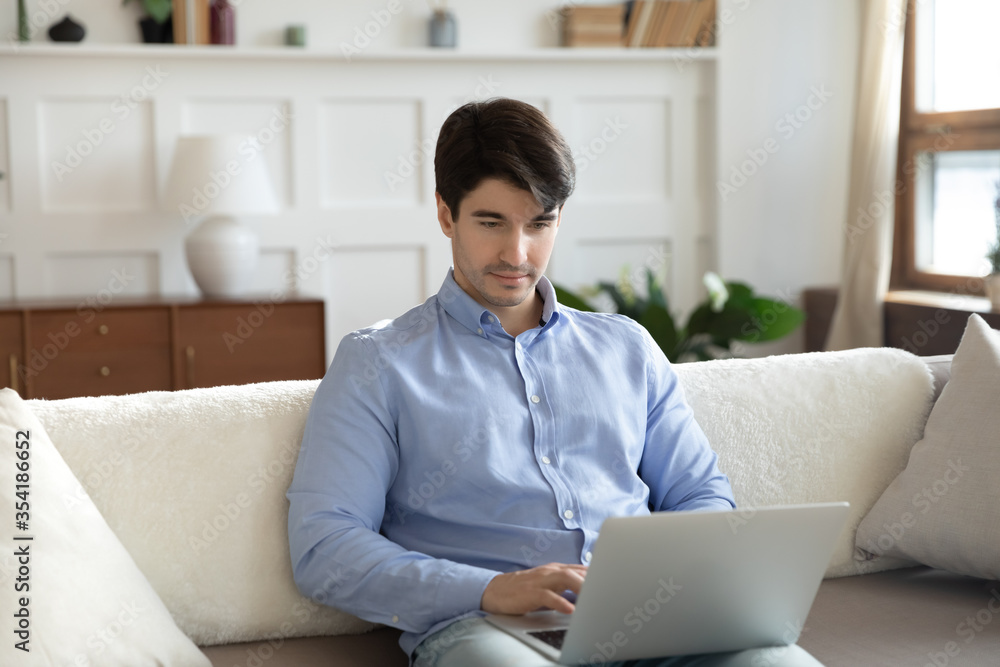 Young caucasian young man sit on sofa in living room working on laptop, typing texting, concentrated millennial male relax on couch at home browsing wireless internet on modern computer gadget Young caucasian young man sit on sofa in living room working on laptop, typing texting, concentrated millennial male relax on couch at home browsing wireless internet on modern computer gadget