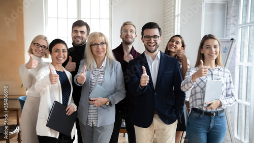 Excited group of diverse different ages businesspeople looking at camera, showing thumbs up gesture. Happy multiracial middle age and young employees evaluating seminar lecture, proposing services.