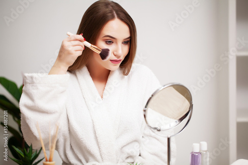 Young woman at home applies makeup on the face in the bathroom in front of a mirror